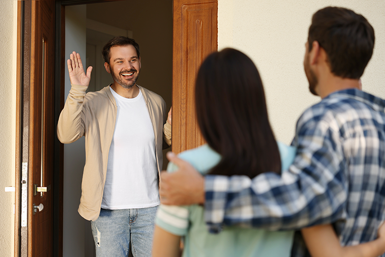 Homme, dans le cadre d'une porte, salue un couple qui se dirige vers lui.